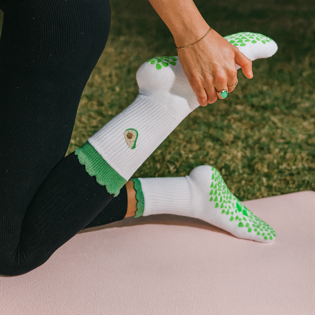 Person stretching outdoors on a fitness mat while wearing white Pilates grip socks with a green avocado design and non-slip traction grips on the sole